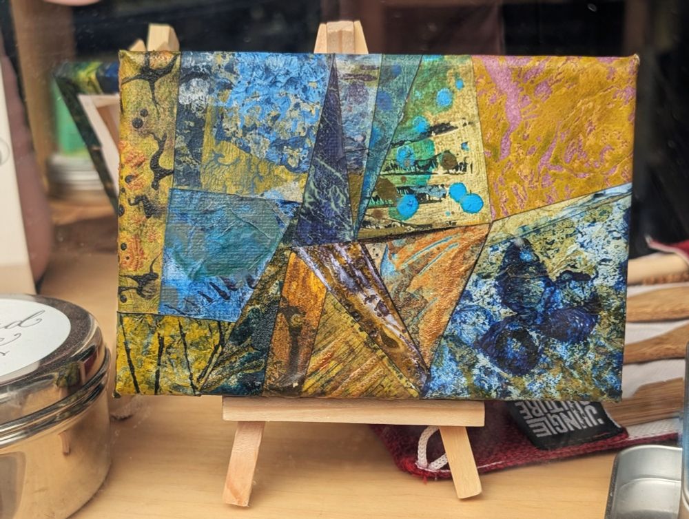 Photograph of a small, mixed media, abstract collage on canvas, featuring organic and geometric textures and patterns in a varied colour palette. The artwork is standing on a mini easel in a shop window.