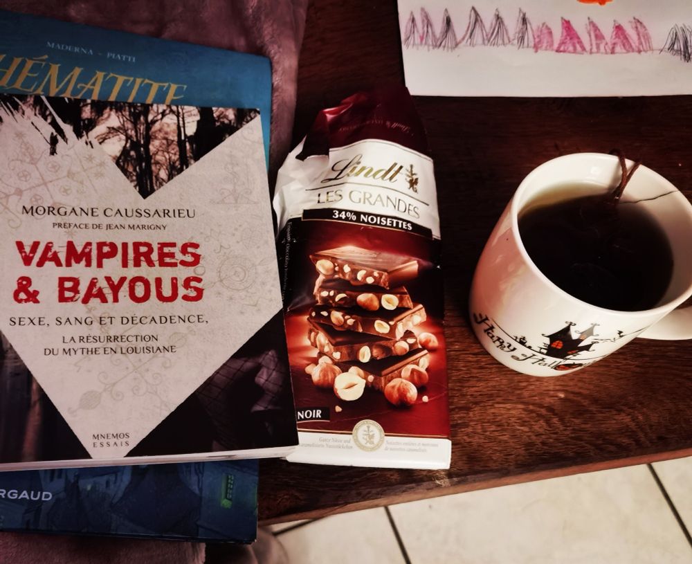 Livre Vampires et bayous de M. Caussarieu et une BD Hematite en dessous, une tablette de chocolat Lindt aux noisettes et une tasse à droite Happy Halloween avec I breuvage de sorcière (aka une tisane)