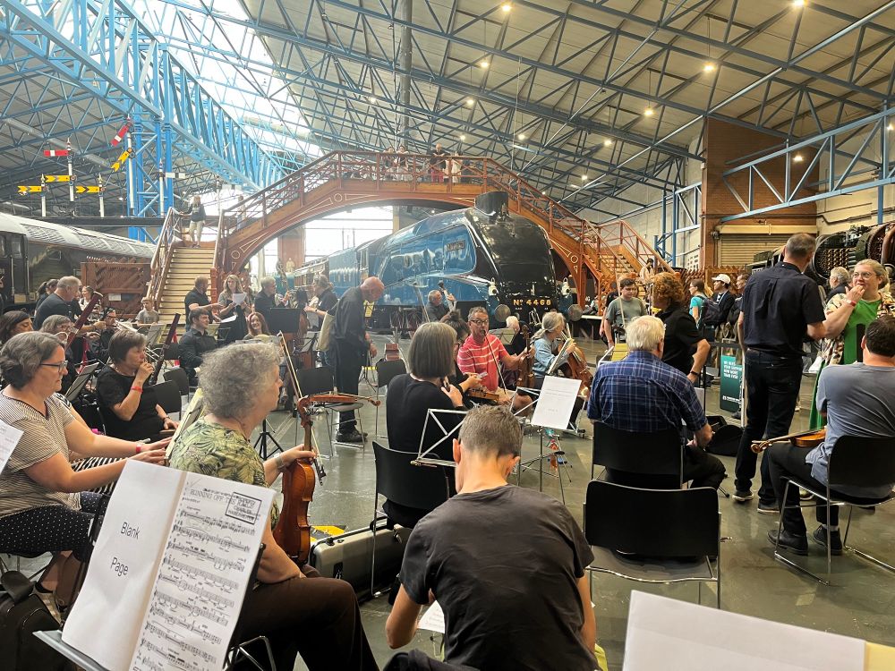 An orchestra, wearing “normal” clothes, setting up in front of The Mallard (fucking epic ace steam engine made in Doncaster 😁) with a slightly bruised general public looking on, including from the wooden bridge over the top of The Mallard 