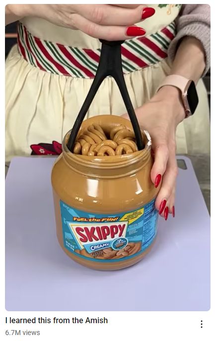 Youtube Shorts preview image of a lady in a pinafore pushing a plastic potato masher into an extra-large jar of Skippy Creamy Peanut Butter, causing the PB to ooze upwards into what looks like a brown nest of snakes.