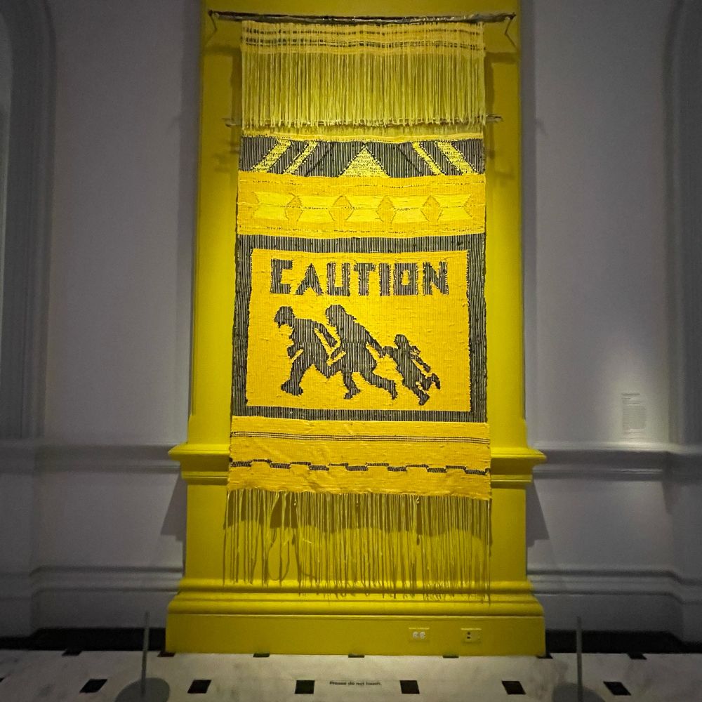 Artist rendition in a bright yellow tapestry of a 1990s California “immigrant crossing” sign depicting a family running with a young child. The title of the piece references the Dick and Jane books.