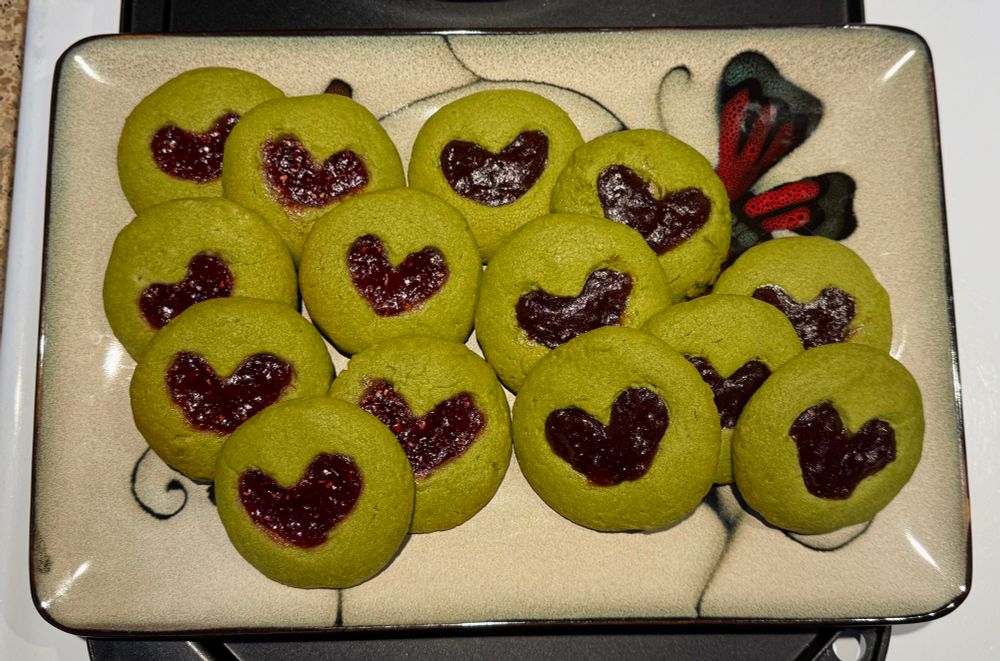 14 freshly baked matcha thumbprint cookies on a serving plate , the thumbprints are heart shaped , half of the cookies have raspberry jam as the center and the other half have apple plum jam 