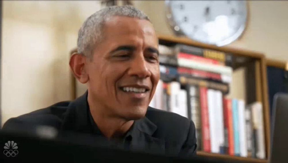 barack obama is smiling while sitting in front of a bookshelf with a clock on it .