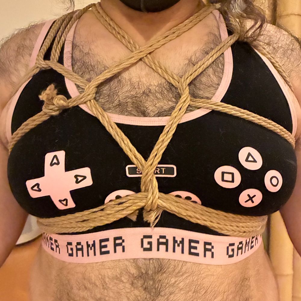 A beautiful chest harness is tied over the chest/breast area of the model. The model is wearing a sports bra style top depicting a video game controller layout. The band at the bottom of the bra says “GAMER” repeatedly. The harness is tied over the bra.
