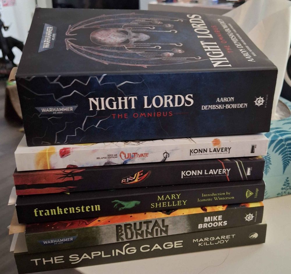 Stack of books. (The Sapling Cage by Margaret Killjoy, Brutal Kunin by Mike Brooks, Frankenstein by Mary Shelly, Rave by Konin Lavery, Cultivate by Konin Lavery, and Night Lords the Omnibus by Aaron Dembski-Bowden). The irony of the post is that the Night Lords omnibus is gigantic compared to the others, but also the one I'm actively reading right now.