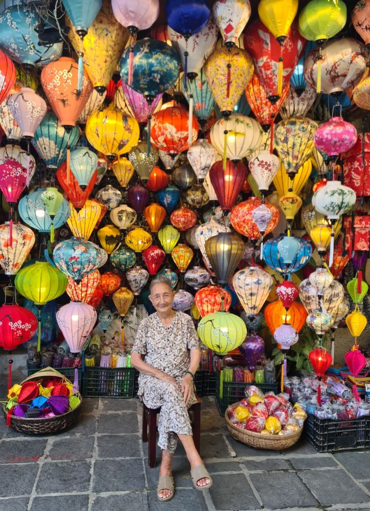 A small smiling lady on a stool surrounded by lots and lots of brightly coloured lanterns of different shapes and sizes

