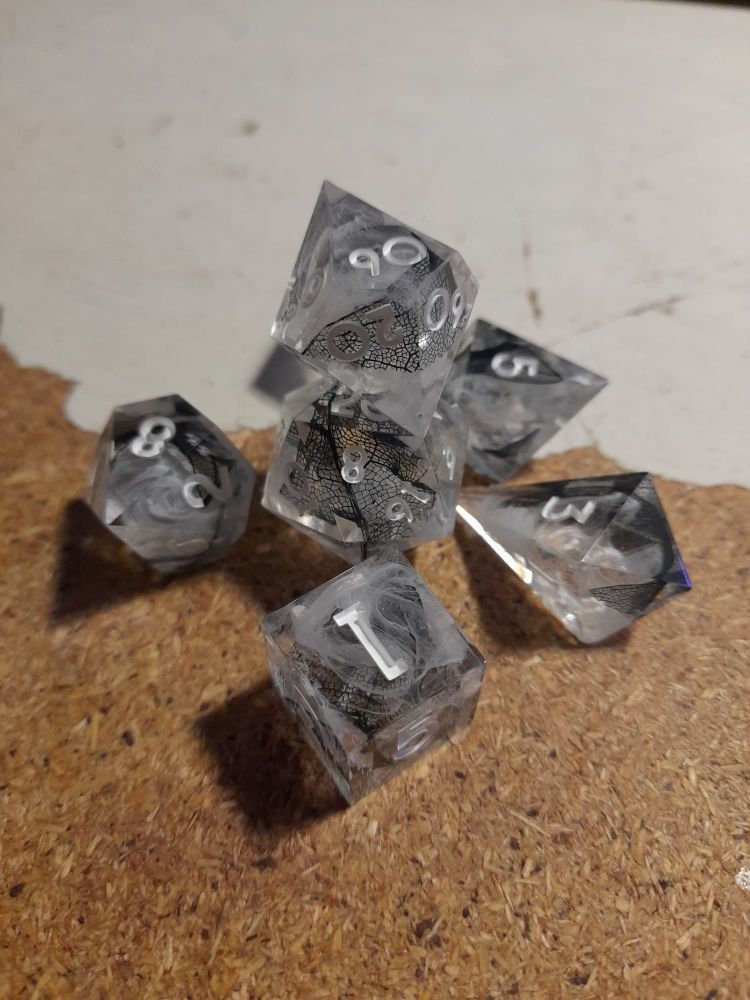 Photo of a set of dice.
The "Of Mist & Memories" set.

Description from the shop:
"A set of 7 ttrpg dice. They have white swirls in clear resin and black skeletal leafs. The numbers are white."
