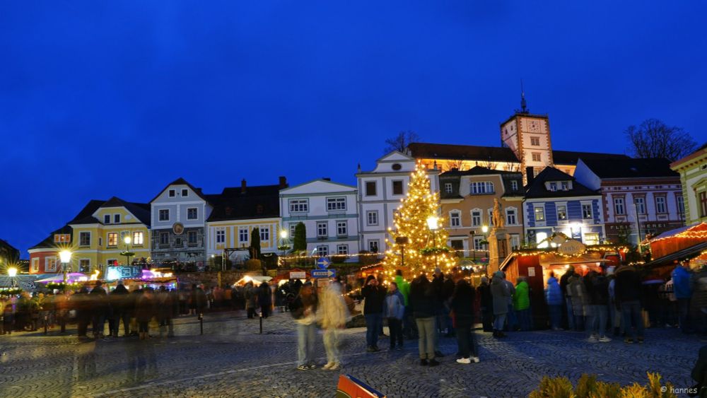 Das Bild zeigt einen festlich beleuchteten Weihnachtsmarkt bei Einbruch der Dunkelheit. Im Vordergrund stehen viele Menschen, die sich um die Verkaufsstände versammelt haben. Die Szenerie ist durch den Kopfsteinpflasterplatz, einen großen, geschmückten Weihnachtsbaum mit Lichtern, und beleuchtete Stände geprägt.

Im Hintergrund befinden sich malerische, bunte Gebäude mit typisch europäischen Fassaden. Weiter oben sieht man einen Uhrturm, der Teil eines Schlosses ist, ebenfalls beleuchtet. Die Atmosphäre wirkt einladend und lebendig, mit weihnachtlicher Dekoration und einem warmen Lichterspiel, das einen Kontrast zum tiefblauen Himmel bildet.

