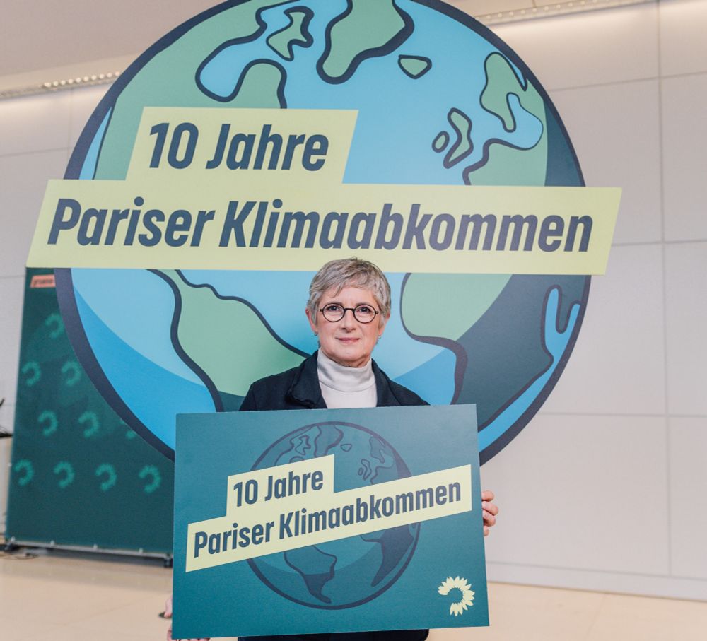 Britta Haßelmann hält ein Schild mit dem Text „10 Jahre Pariser Klimaabkommen“ hoch. Dahinter ist eine Weltkugel zu sehen.
