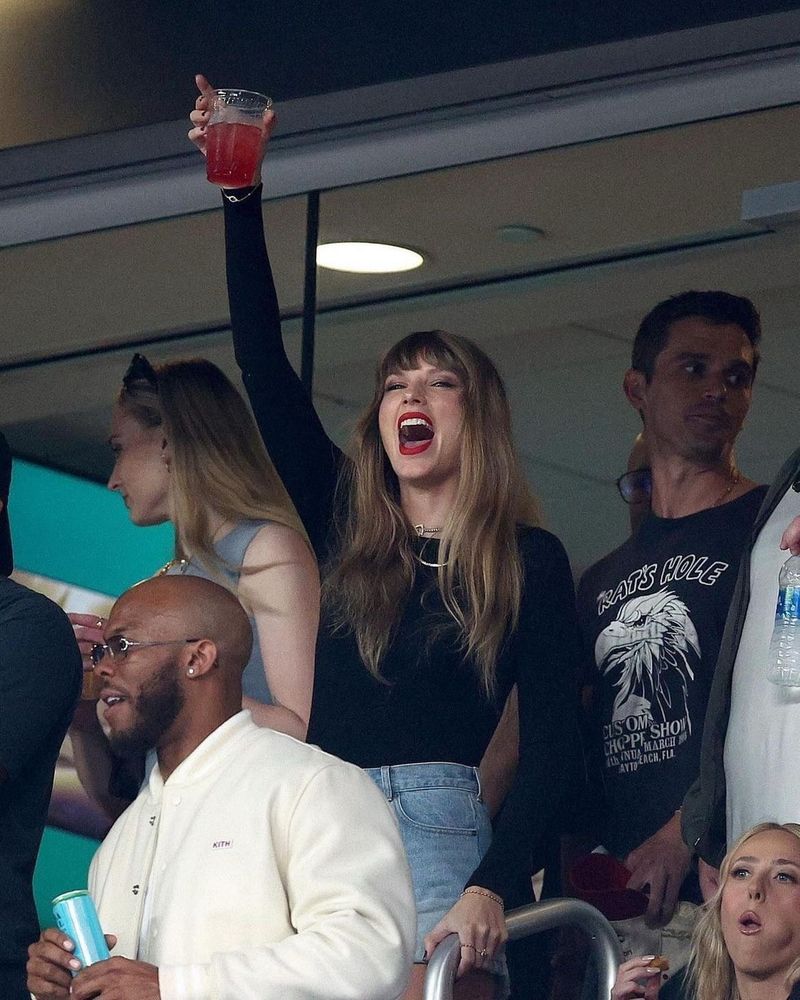 Taylor Swift in the stands at a football game, joyfully holding up a beverage