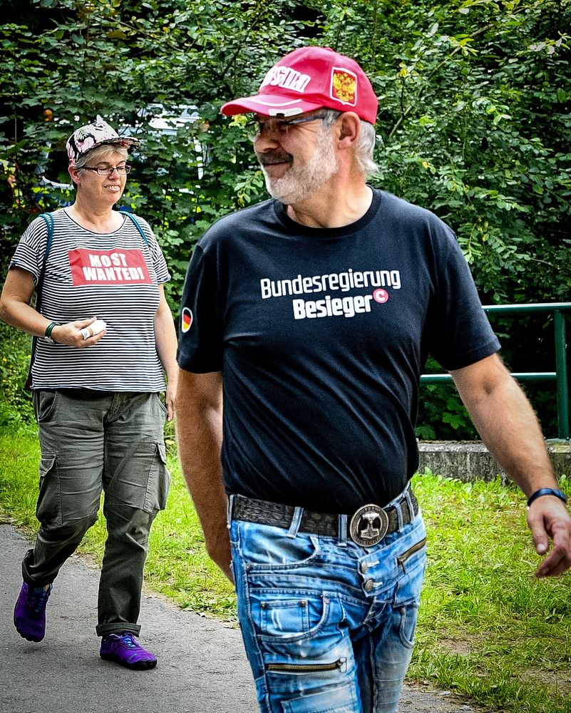 Ein Teilnehmer trägt ein Shirt „Bundesregierung Besieger“ mit Chen Compact-Logo. Dahinter eine Frau mit „Most Wanted“ auf dem Shirt. 