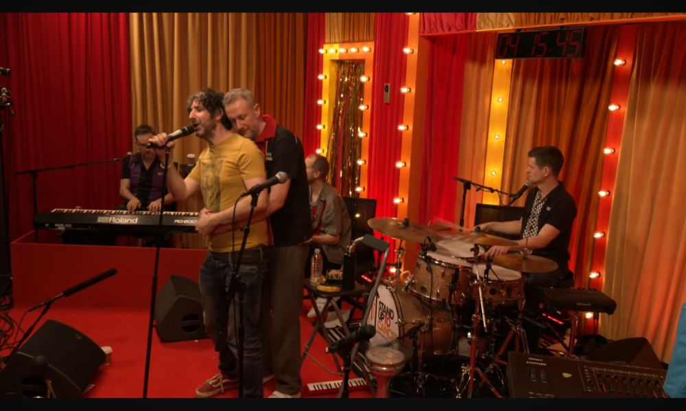 A screenshot from The Horne Section's 24 hour challenge playing a single song constantly.  Mark Watson is singing into a microphone on a stand and Alex Horne is standing behind Mark with his arms wrapped around his middle and resting his head on Mark's shoulder.  
Ben Reynolds is playing the drums behind them and on their right are Will Collier on the bass guitar (although we can't see his guitar, obscured behind Mark and Alex) and Ed Sheldrake on the keyboard.  The digital clock indicating how long they'd been going at that point says 4hrs, 15mins, 45secs.