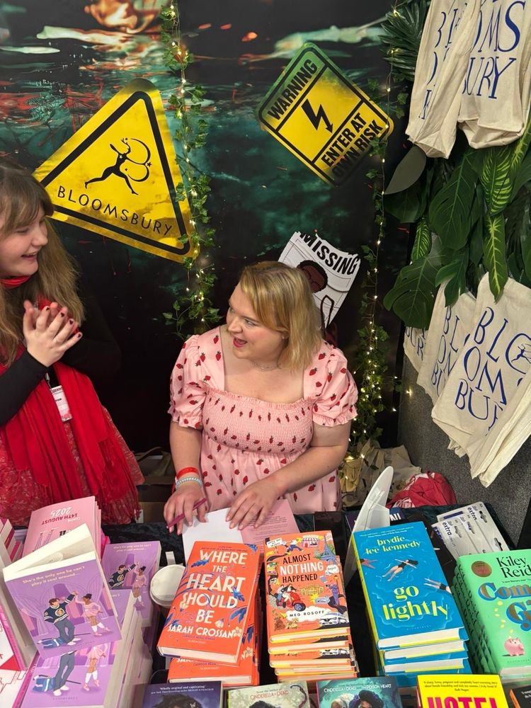 Photo of a woman with short blonde hair wearing a pink strawberry print dress, sitting behind a table full of books. She’s smiling and talking to someone next to her while signing a booklet in front of her.
