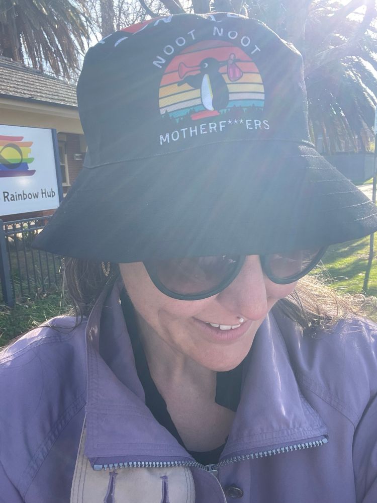 Close up selfie of a basic white femme wearing sunnies and a bucket hat - head is tipped down to emphasise the hat which is black with a graphic of Pingu holding a bindle and tooting their beak and text around it saying ‘Noot Noot Motherf***ers’
