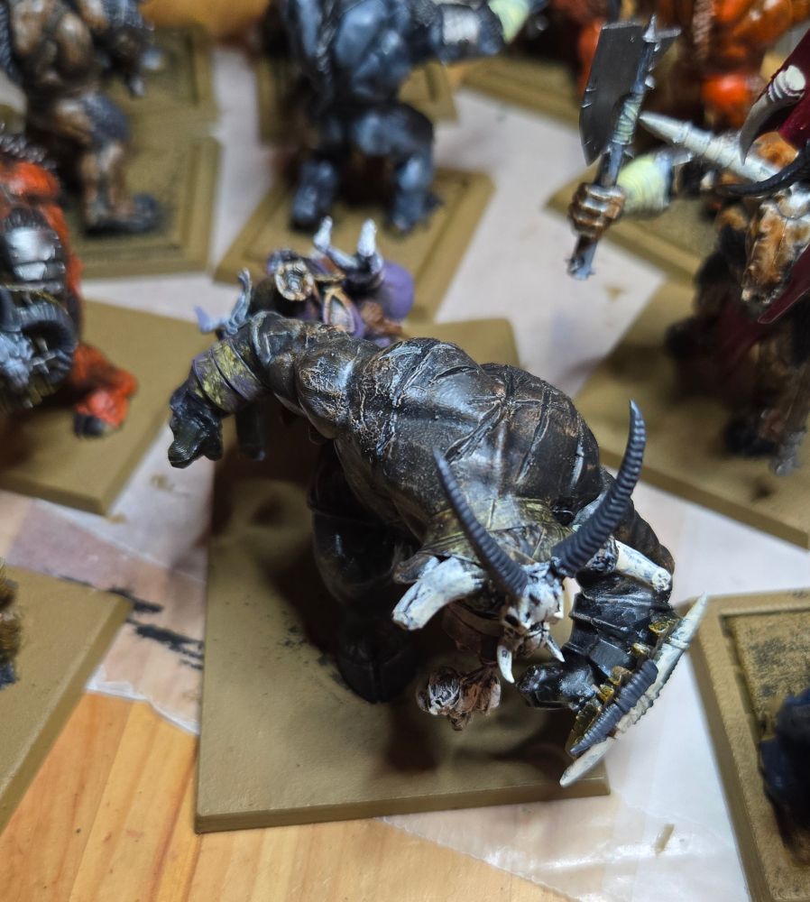 A Minotaur pulling the near wagon from another angle. They have a shield on their front left arm and their horns are broken. A demonic goat skull has been nailed to their forehead. 