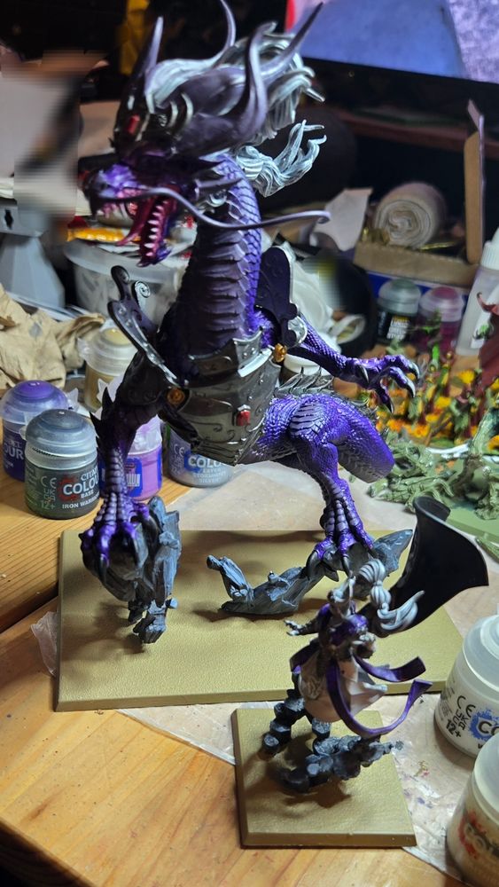 A now fully constructed and further painted Miao Ying the Storm Dragon model sits on a coffee table. The bases have been painted the colour of dried mud, the two feature a predominantly purple motif and white hair with Grey undertones. 