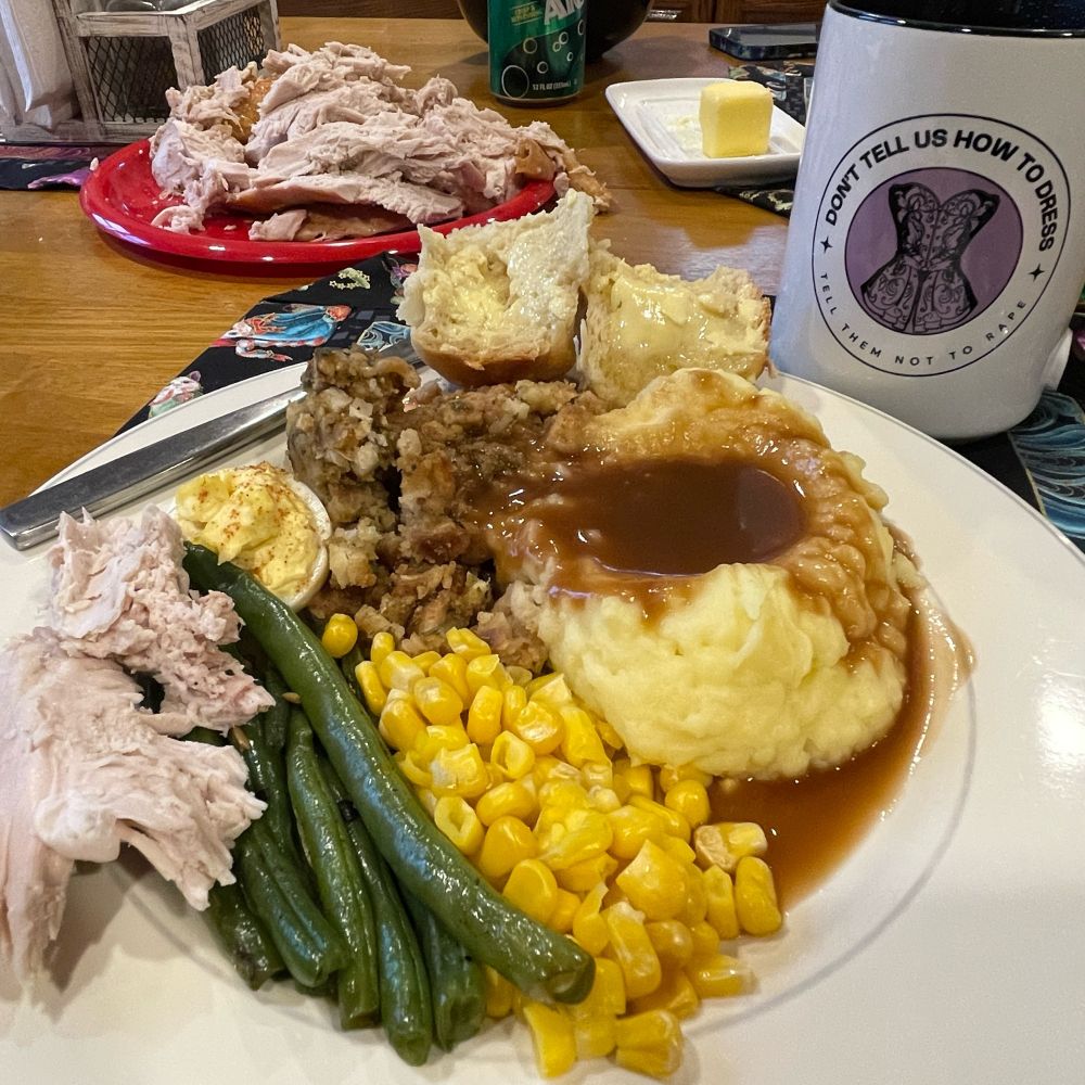 Plate of Thanksgiving food including turkey, corn, mashed potatoes, stuffing, rolls, and gray. Mug in background reads “don’t tell us how to dress, tell them not to rape.”