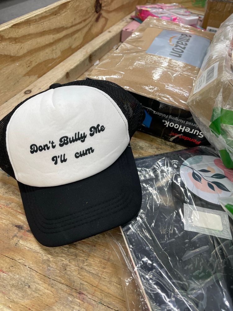 Black and white hat sits in wooden oversized bin along with several other unopened boxes. Hat reads “Don’t Bully Me I’m Cum.” 