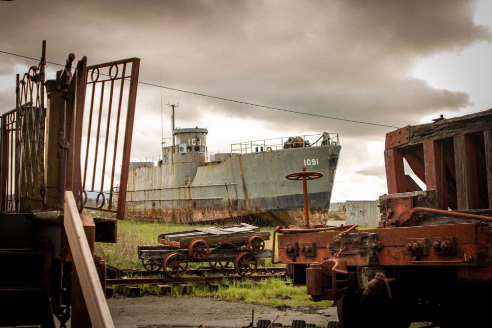 A photo of an old ship that is now on land and is rusting. In the foreground there are various train parts and a stormy sky that sets the mood. 