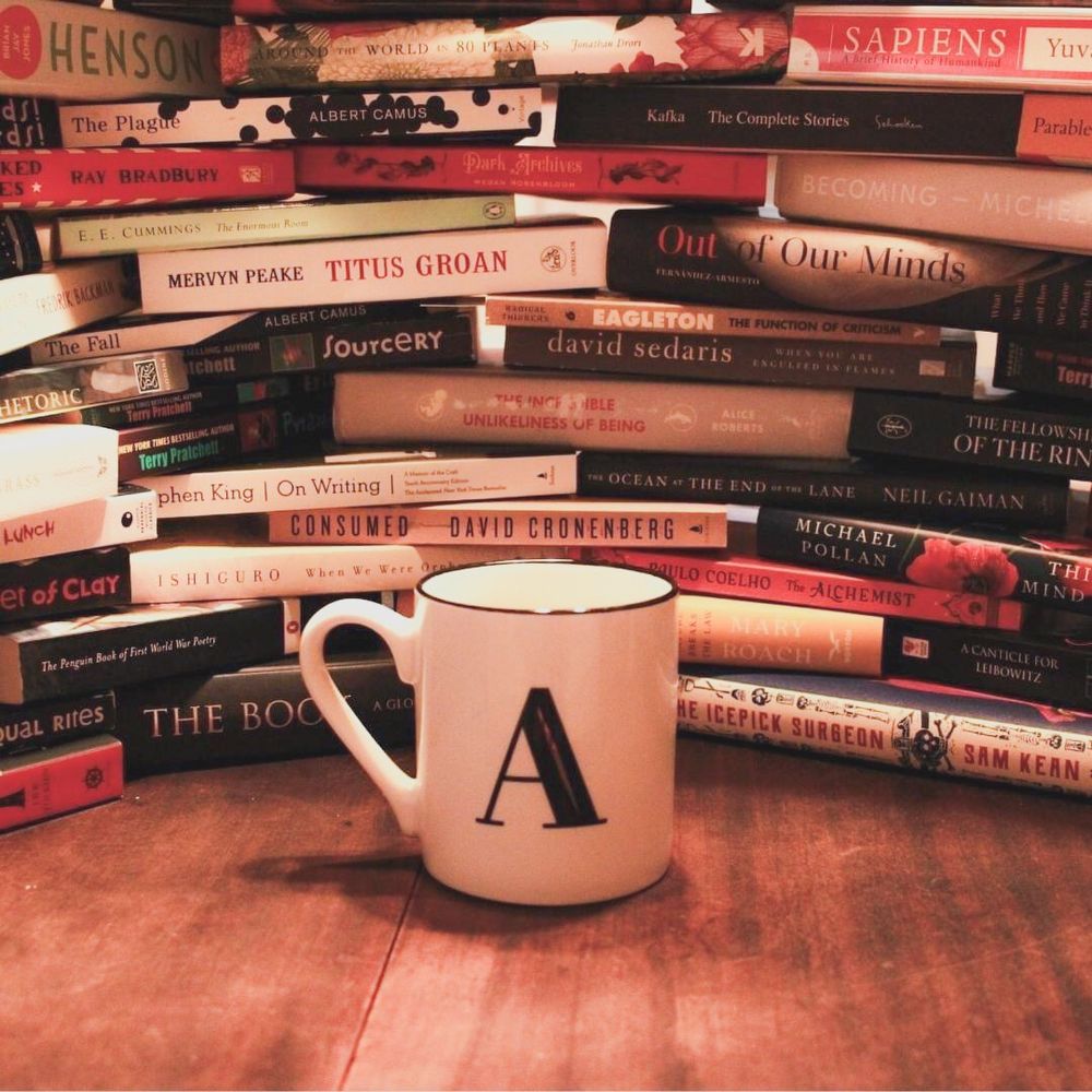 A photo of many books stacked on top of each other on an old wooden table top. A white mug with the capital letter A is placed in front of the books. 