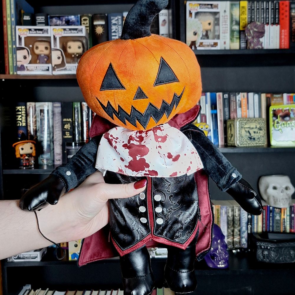 Hand holding up a headless horseman plush with a jackolantern for a head. Bookshelves are in the background.