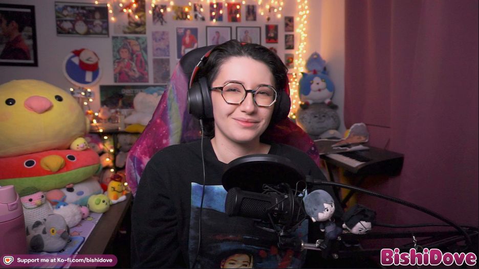 Bishi smiles at the camera in their stream room while wearing a black long sleeved shirt with a graphic design on it 