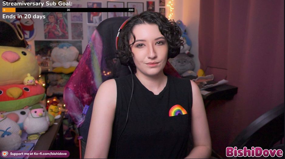 Bishi smiles at the camera in their stream room while wearing a black sleeveless shirt with a rainbow on it