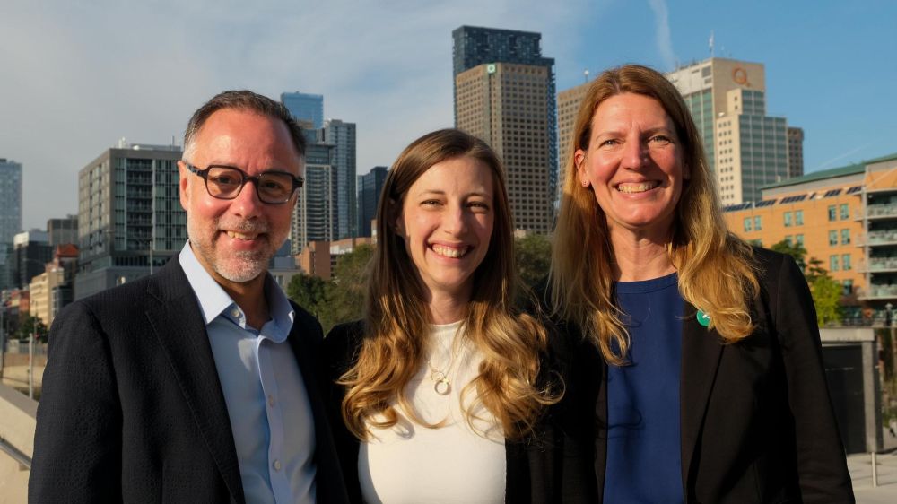 De gauche à droite : Luc Rabouin, Élise Tanguay et Véronique Fournier posent ensemble dehors, souriants, avec les immeubles du centre-ville de Montréal derrière eux.