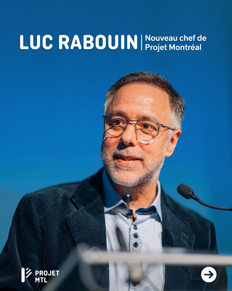 Luc Rabouin, portant un veston bleu, parle devant un micro sur fond bleu avec un texte en haut indiquant "Luc Rabouin | Nouveau chef de Projet Montréal". Le logo de Projet Montréal est en bas à gauche.