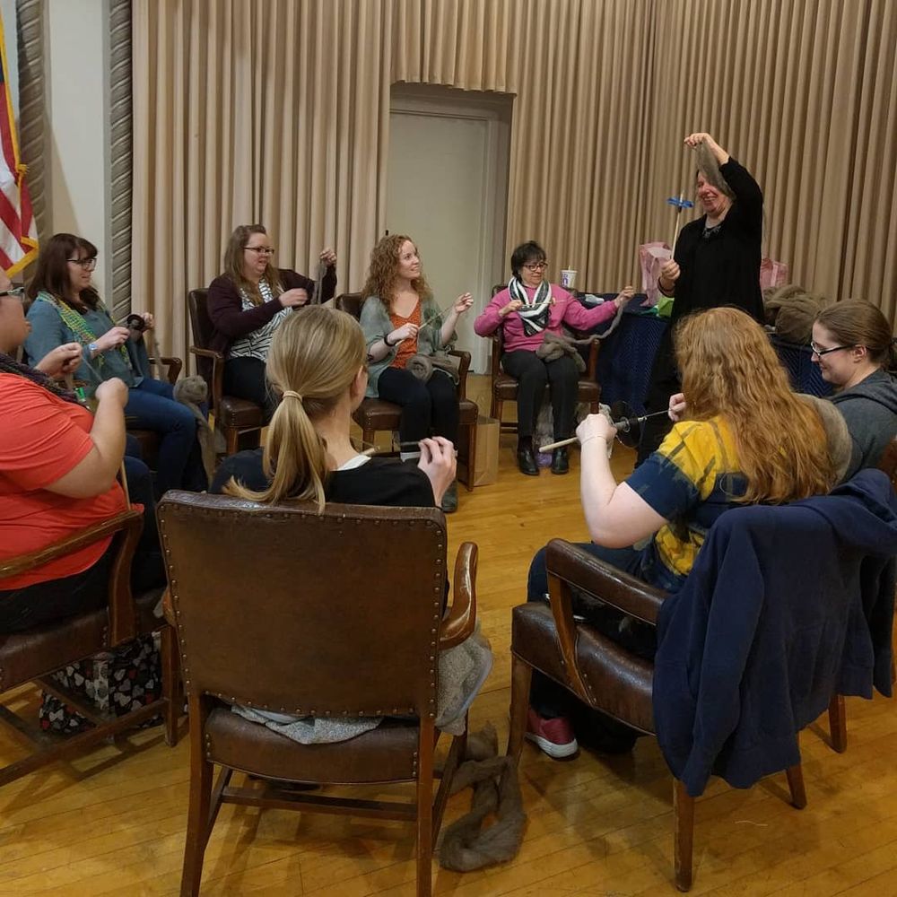 a handspinning workshop with students sitting in chairs holding drop spindles with instructor standing and demonstrating the technique