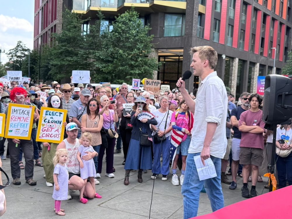 Young man with short blond hair speaking into a microphone, with the audience in the background. In the foreground, near the speaker, are two small girls in identical striped dresses. One is gazing at the speaker, open-mouthed.