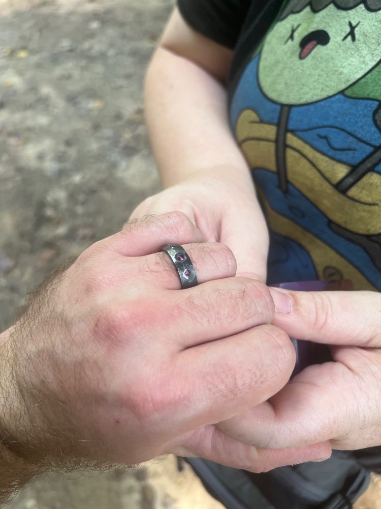 Jackie’s hands holding my left hand, which bears the engagement ring she proposed to me with. It’s sterling silver that looks like dark hammered iron with three rubies inlaid in it, the largest of which is in the center.