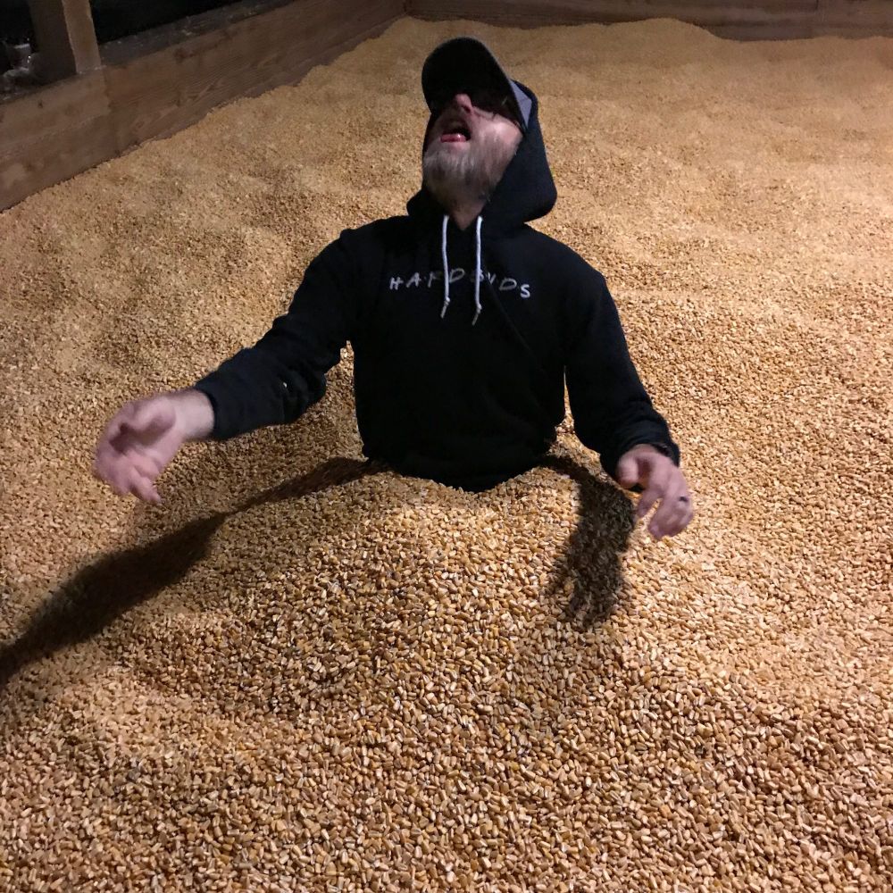 Tom sitting, covered to his waist, in a pit of loose corn kernels. His head his tipped back - he is about to catch a piece of corn in his mouth. Children have been playing in this corn all day. The whole story here is gross