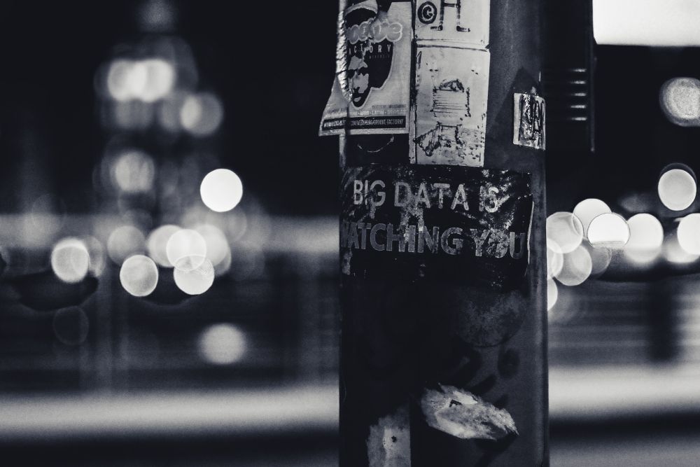 Black and white photo of a sticker on a lamp post that reads "big data is watching you." Photo by ev on Unsplash