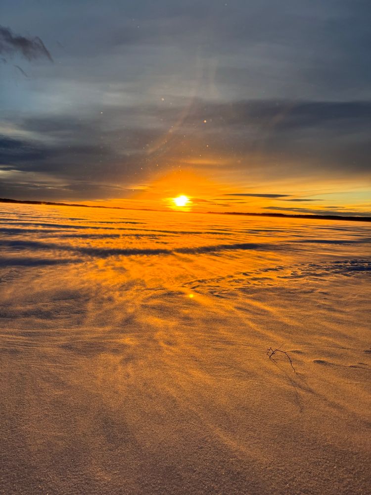 Kuva järven jäältä. Aurinko paistaa pilviverhon ja lumituiskun takaa. 