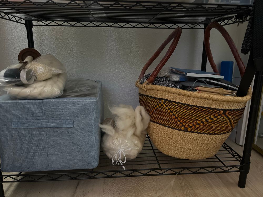 a shelf with a bin and a basket on it. squeezed in between the bin and the basket is a bag of fiber and a drop spindle