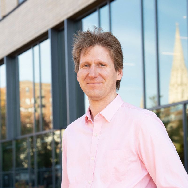 Christian Kästner, Associate Professor at Carnegie Mellon University, smiling at the camera while wearing a light pink button-down shirt. He stands in front of a modern glass building with a church spire visible in the background cityscape