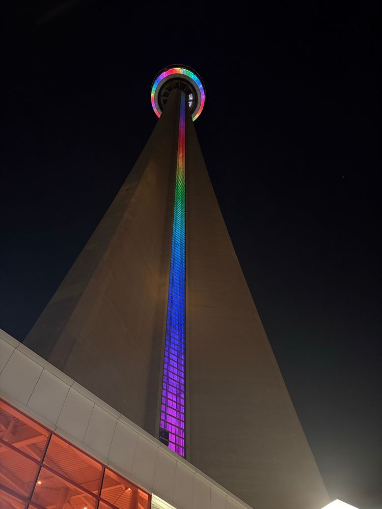 The CN tower taken from a low angle. The elevator shaft and little circular part at the top are glowing rainbow colours 
