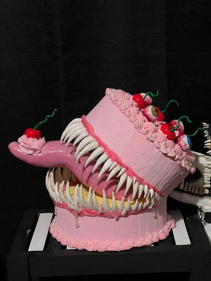 A monster birthday cake in pink icing with a pink tongue holding a cherry and cherry eyeballs and teeth