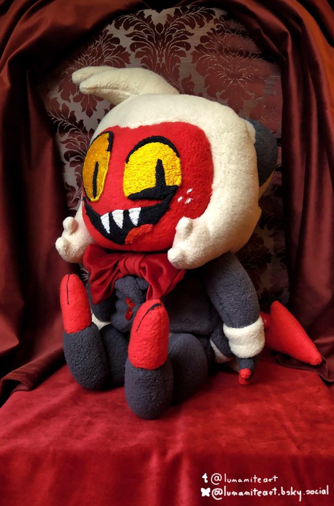 This is a full-body picture of handmade plushie of Moxxie from Helluva Boss. The imp has a big head, white hair, black and white horns. He's wearing a grey tuxedo with a big red bow. The photo of the plushie is taken on an elegant red patterned background with deep-dark red curtains. 