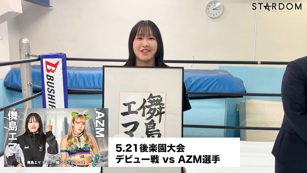 STARDOM trainee Ema Maishima holding up the frame with her ring name "舞島エマ" written inside towards the camera. A graphic for her debut match against AZM is on the bottom-right of the screen. They are in the STARDOM dojo with President Taro Okada.
