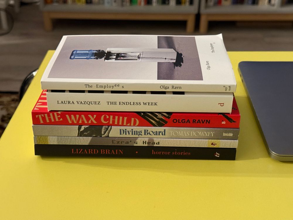 Stack of books: Olga Ravn “The Employees”; Laura Vazquez “The Endless Week”; Olga Ravn “The Wax Child”; Tomas Downey “The Diving Board”; David Kuhnlein “Ezra’s Head”; “Lizard Brain: Horror Stories”