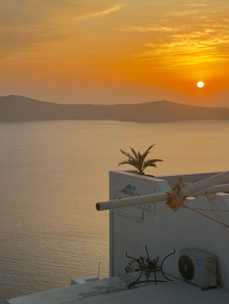 A bright orange and yellow sunset, viewed from a whitewashed building with a flat roof. The building sits above a body of water.There are undulating mountains silhouetted on  the other side of the water.