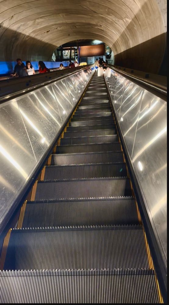 Descending on a long escalator. Ridged metal stairs are sidelit. It is a very long escalator.