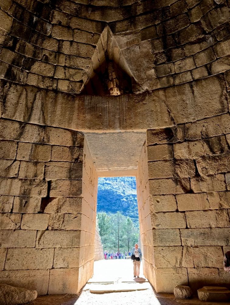 The door of this honeycomb like structure, constructed of brown rock, opens to a view of the mountainous terrain of the Peloponnese. A person stands sideways in the doorway.