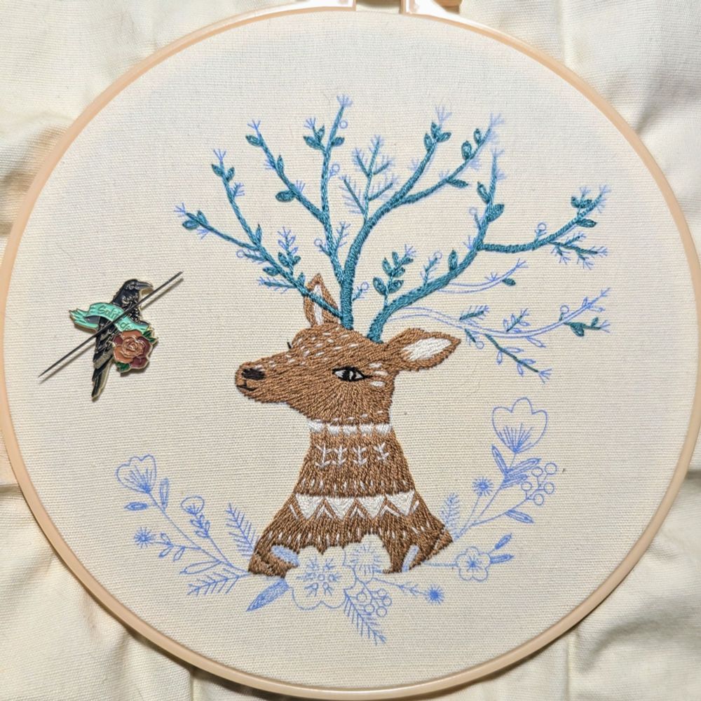 An in progress embroidery piece on cream cloth. A folk art style deer stitched in brown with white stylistic markings, from the chest up. The antlers are blooming branches, partially finished, in dark teal thread