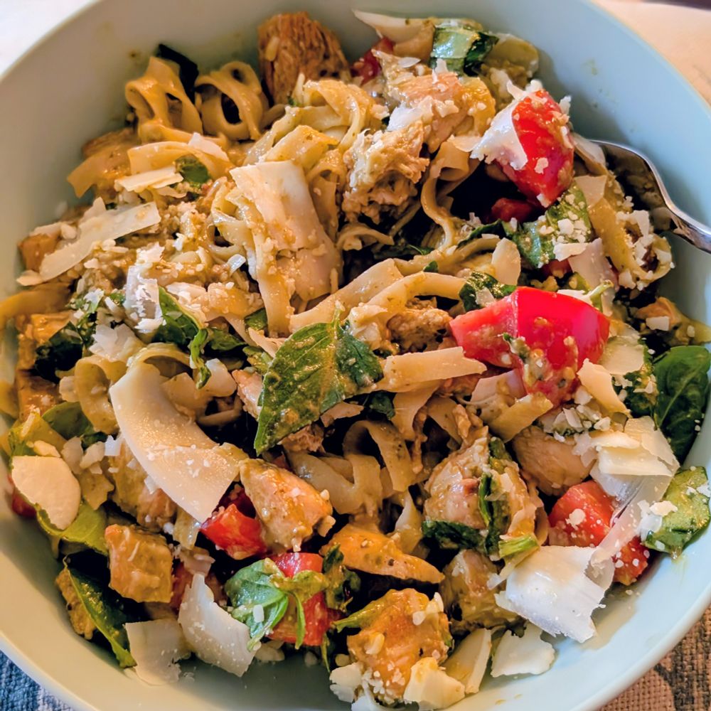 A bowl of long flat pasta mixed with pesto, Basil leaves, chicken, chopped tomatoes and shaved Parmesan 