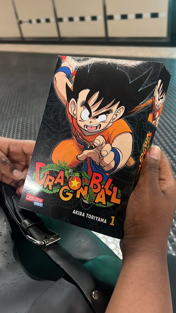 Eine Person of Colour hält den ersten Band des Mangas „Dragon Ball“ von Akira Toriyama in den Händen. Auf dem farbigen Cover ist der junge Protagonist Son-Goku in orangefarbener Kleidung mit einem blauen Gürtel und Armbändern abgebildet. Er streckt entschlossen eine Faust nach vorne. Der Titel „Dragon Ball“ ist bunt gestaltet, mit einem grünen Drachen, der sich um die Buchstaben windet. Unten auf dem Cover steht „Carlsen Manga!“ und „Akira Toriyama 1“. Im Hintergrund ist ein U-Bahn-Bahnsteig mit Fliesenboden und Wänden im Retro-Stil zu sehen. Ein schwarzer Rucksack mit Metallschnalle liegt auf dem Schoß der Person.
