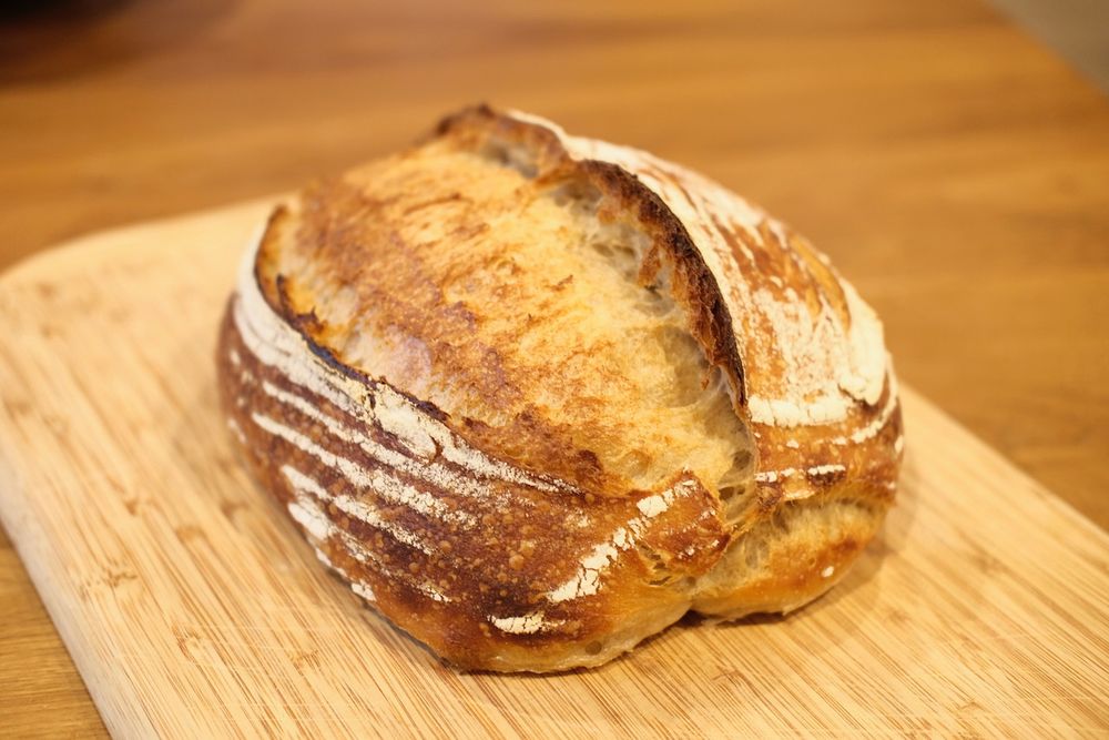 Fresh-baked sourdough batard sitting on a wooden cutting board on a walnut wood table.