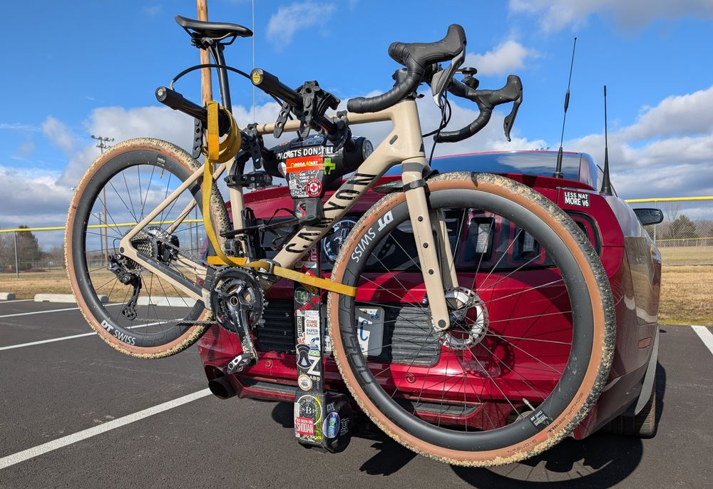A slightly muddy gravel bike on a sticker-covered bike rack attached to a red mustang with antennas 